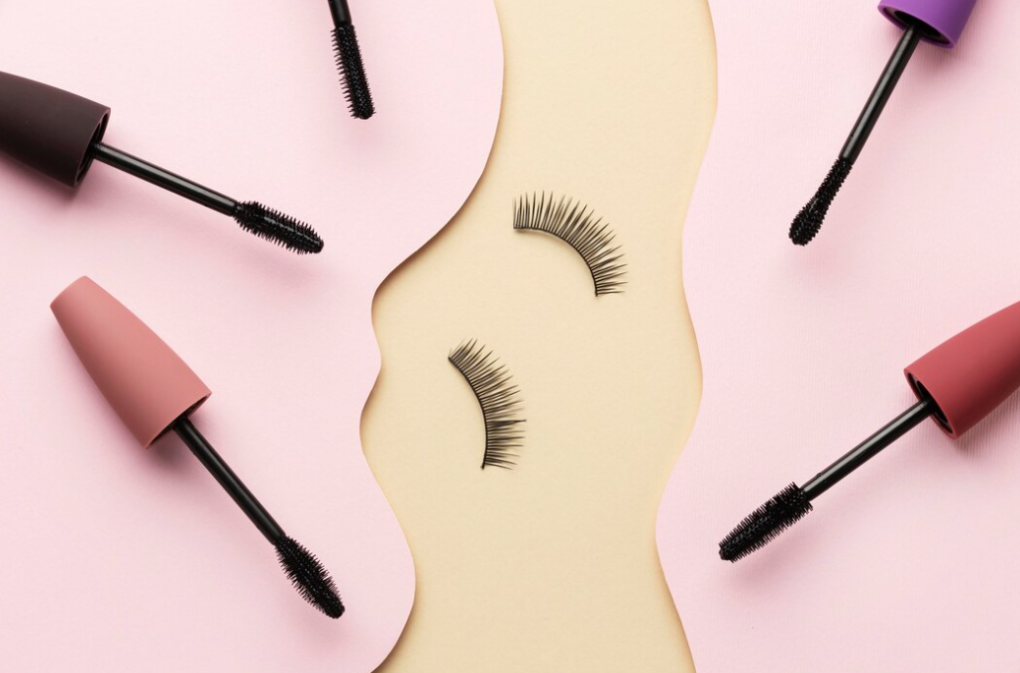 Flat lay of mascara wands and false eyelashes on a pink background with a wavy beige divider.