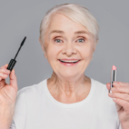 old woman smiling and holding a mascara and lipstick in front of a gray wall