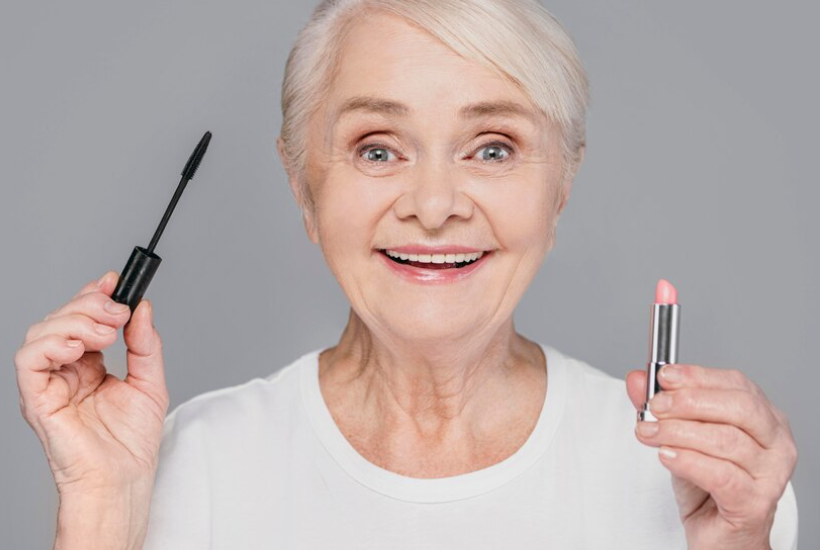 old woman smiling and holding a mascara and lipstick in front of a gray wall